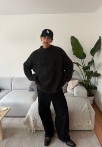 Pull en maille épaisse et sombre avec un col montant, pantalon noir à jambes larges, et chaussures en cuir noir. En arrière-plan, une plante et un canapé clair.
