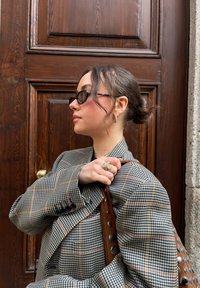 Blazer oversize a motivo houndstooth grigio e marrone con accenti arancioni. Borsa marrone con hardware circolare. Occhiali da sole neri. Orecchini a cerchio in argento.