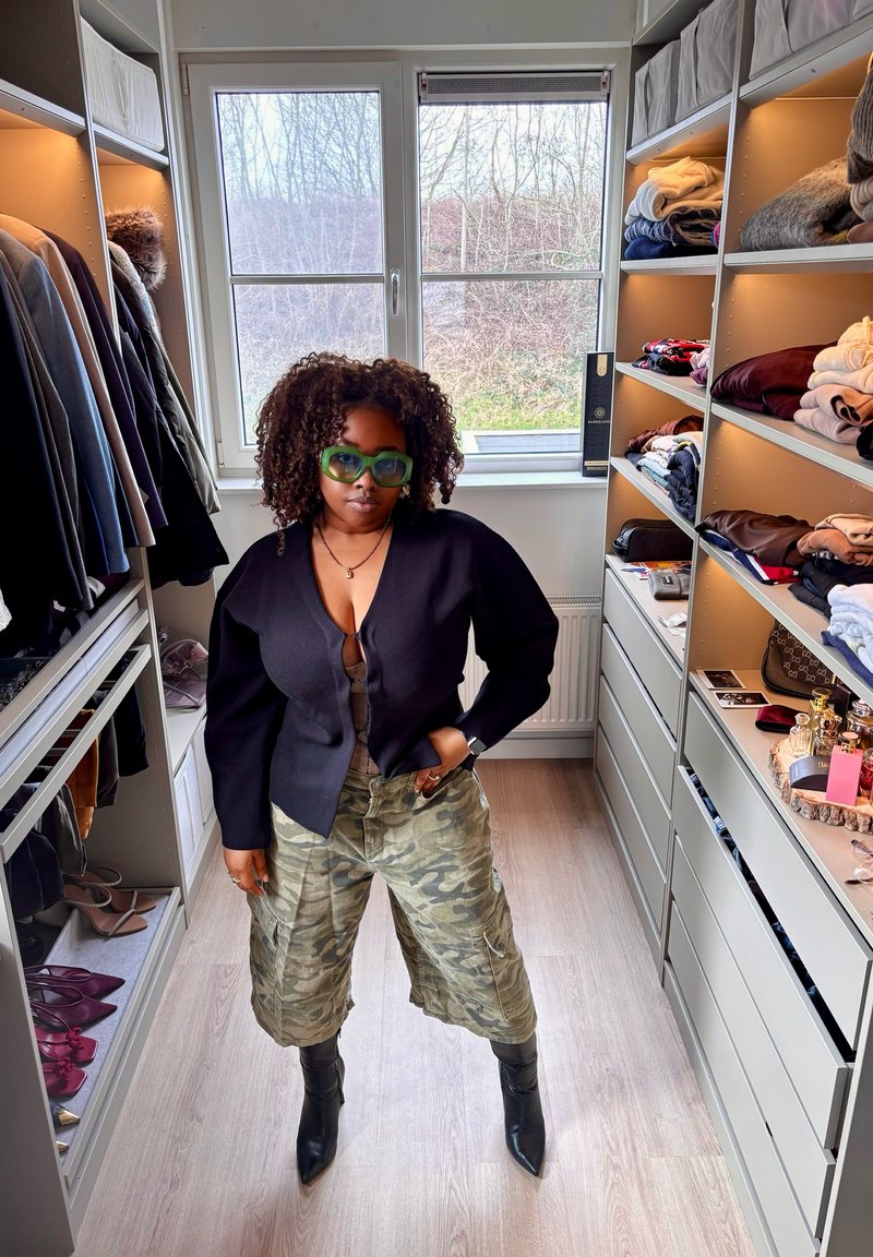 Kvinde med krøllet hår og grønne briller står i et velorganiseret gå-i-closet iført sort top, camouflage vidde bukser og sorte støvler.