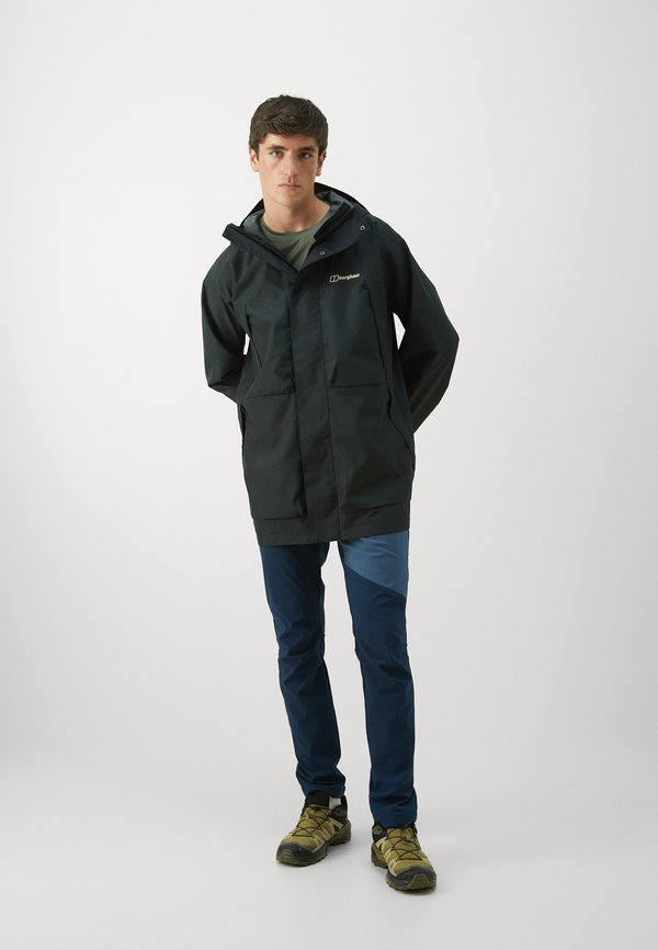 Giacca nera impermeabile con cappuccio, dotata di due tasche frontali e un logo. Indossata sopra pantaloni blu e verde acqua a blocchi di colore e scarpe da escursionismo.