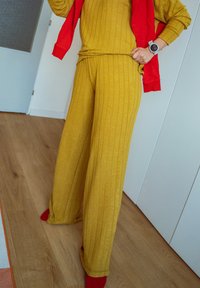 Personne portant un pantalon et un haut en tricot côtelé jaune moutarde avec des chaussettes rouges, tenant une veste rouge, debout sur un sol en bois à l'intérieur.