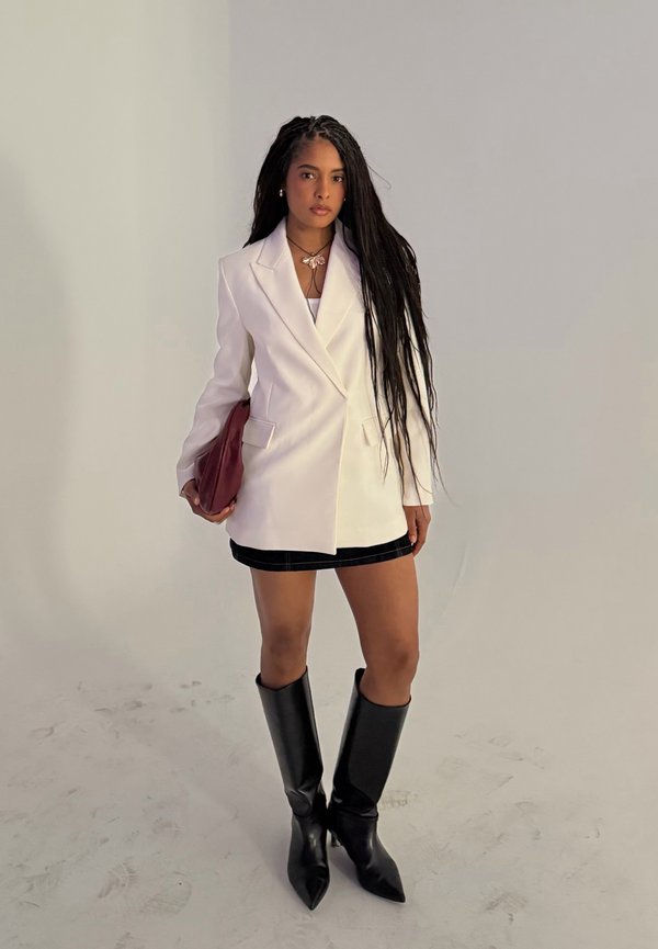 Vrouw met lang haar, gekleed in een witte blazer, zwarte minirok, zwarte kniehoge laarzen, die een rode clutch vasthoudt, staand tegen een effen achtergrond.