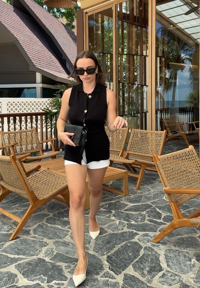 Top sin mangas negro con detalles de botones dorados, pantalones cortos blancos y tacones de punta blanca. Suelo con patrón de piedra y asientos exteriores de madera.