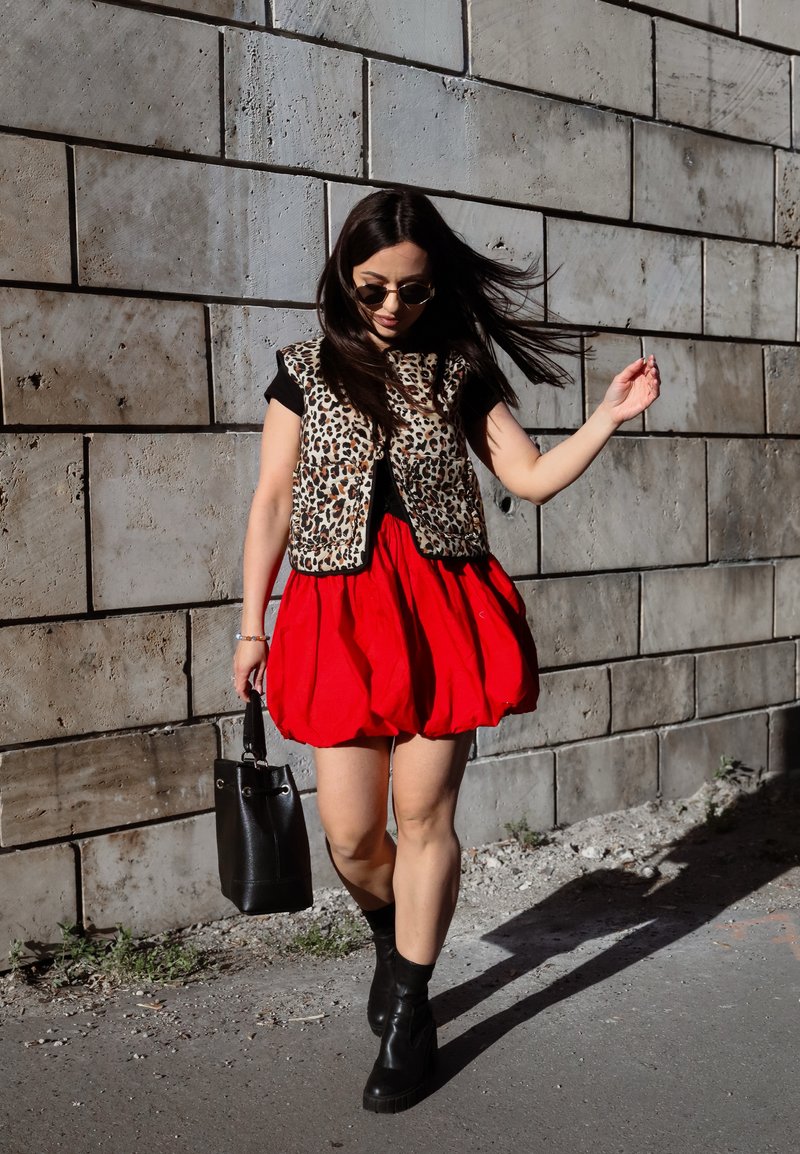 Femme marchant le long d'un mur en pierre, portant un gilet imprimé léopard, une jupe rouge, des bottes noires, des lunettes de soleil et un sac à main noir.