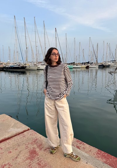 Camisa de rayas de manga larga en marrón y blanco, pantalones anchos color crema y zapatillas verdes, de pie junto a un puerto con veleros. El agua tranquila refleja el paisaje.