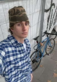 Une chemise à carreaux bleue, un T-shirt blanc et un bonnet camouflage portés par une personne près d'un vélo bleu avec des accents noirs et un antivol visible.