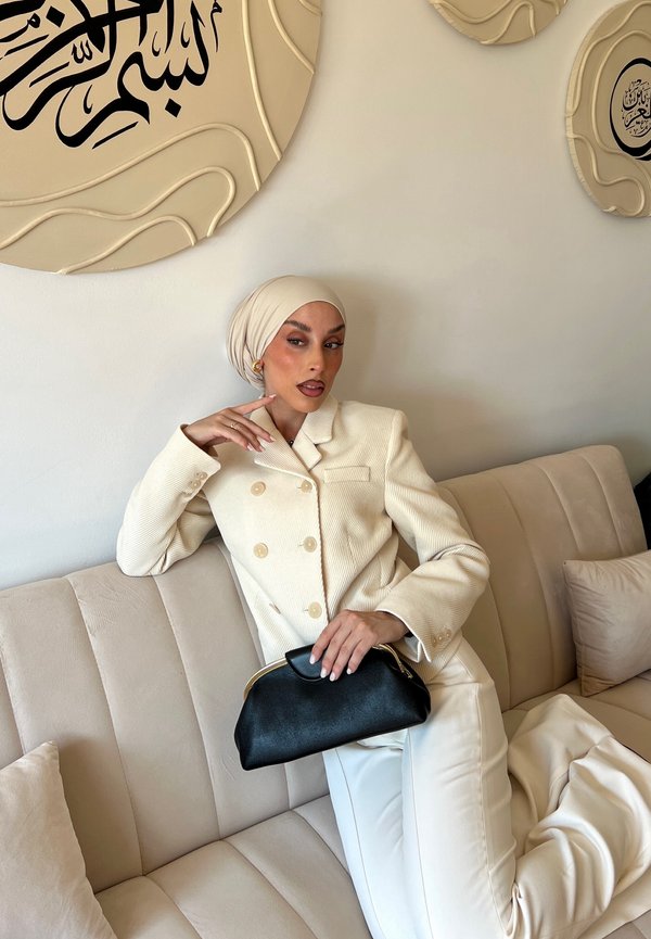 Woman wearing cream blazer and hijab reclining on beige couch, holding black purse, with abstract Arabic calligraphy wall art behind her.