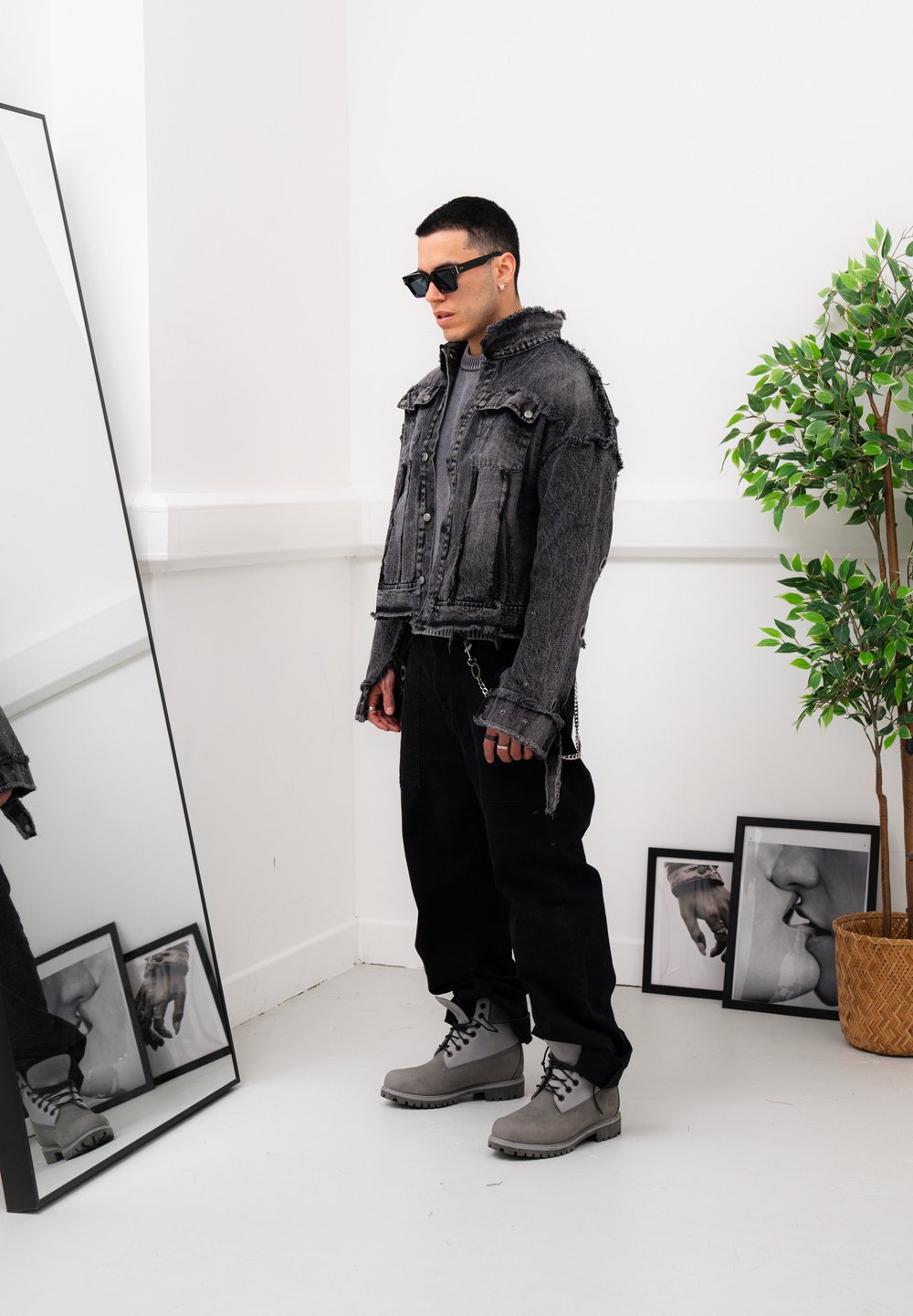 Black denim cropped jacket with frayed edges, black cargo trousers, and grey lace-up boots. The look is accessorised with sunglasses and a metallic chain detail.