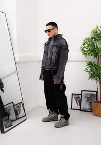 Black denim cropped jacket with frayed edges, black cargo trousers, and grey lace-up boots. The look is accessorised with sunglasses and a metallic chain detail.