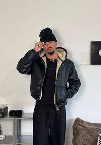 Schwarze Leder-Bomberjacke mit einem cremefarbenen Fleece-Futter, kombiniert mit einem schwarzen Henley-Shirt und schwarzen Hosen, getragen mit einer schwarzen Mütze.