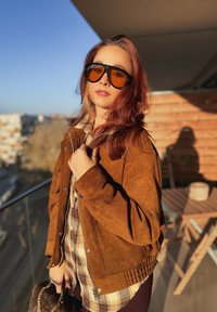 Giacca in camoscio marrone con orlo a coste, abbinata a una camicia a quadri. Grandi occhiali da sole neri con lenti arancioni, in piedi su un balcone.