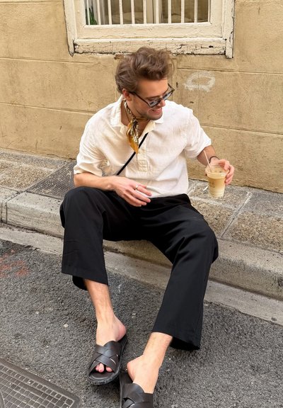 Joven con gafas, camisa blanca, pantalones negros y sandalias sentado en el bordillo de la calle sosteniendo un café con hielo y sonriendo mientras mira hacia un lado.