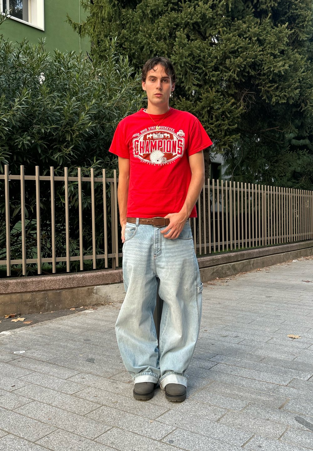 T-shirt rouge avec l'impression "CHAMPIONS", jean léger bleu clair ample, chaussures noires, debout en plein air devant un feuillage vert et une clôture.