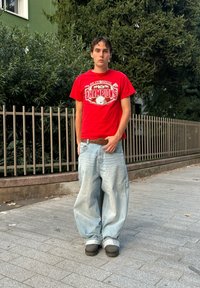 T-shirt rouge avec l'impression "CHAMPIONS", jean léger bleu clair ample, chaussures noires, debout en plein air devant un feuillage vert et une clôture.