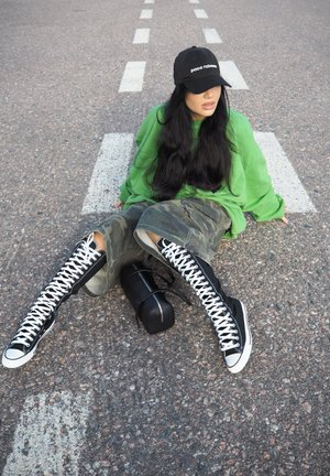 Botas altas negras con cordones blancos, sudadera verde de gran tamaño y pantalones cortos de camuflaje. Gorra negra con logo y pequeña bolsa negra en el suelo.