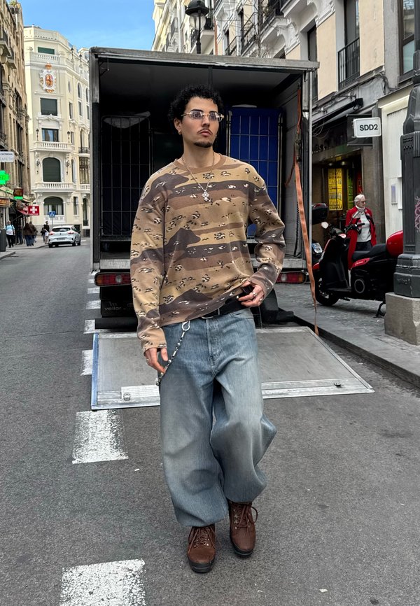 Man met krullend haar die een zonnebril draagt, camouflage trui, wijdvallende spijkerbroek en bruine laarzen staat op een stadsstraat voor een open vrachtwagen.