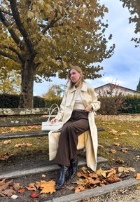 Trench coat creme sobre um sweater bege, calças castanhas de cintura alta, botas pretas de bico fino e uma mala cinza clara, sentada num banco de parque rodeada de folhas de outono.