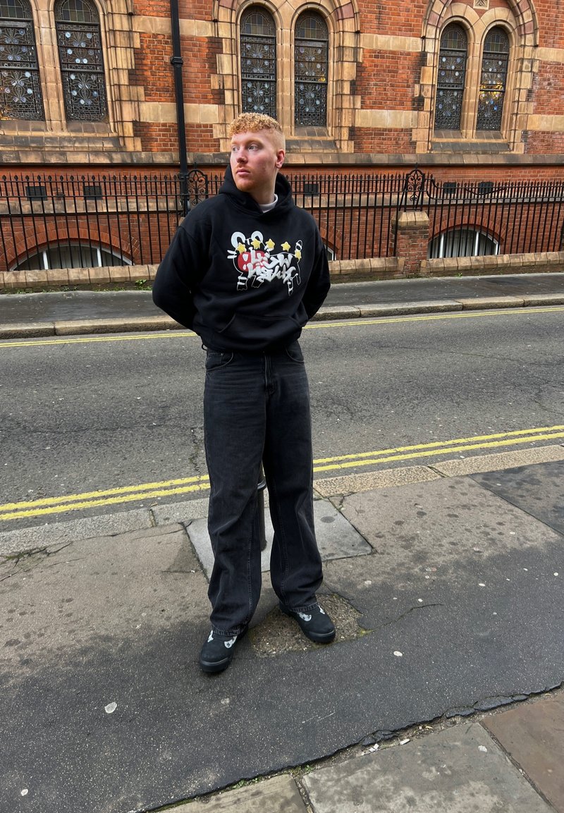 Μαύρη κουκούλα με γραφικό σχέδιο, ξεθωριασμένα μαύρα φαρδιά τζιν και μαύρα παπούτσια. Το υπόβαθρο έχει κόκκινους τοίχους από τούβλα και μεγάλες παράθυρα.