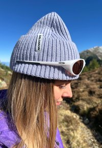 Donna con giacca viola indossa un berretto di maglia grigio della Salomon con occhiali da sole bianchi poggiati sopra, all'aperto in un paesaggio montano soleggiato.