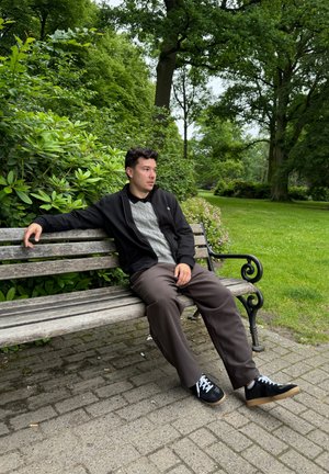 Chaqueta negra con cremallera, suéter gris de punto trenzado, pantalones marrones, zapatillas negras con cordones blancos, sentado en un banco de madera en un parque verde.