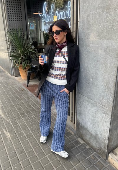 Mujer con gafas de sol, pantalones estampados y chaqueta, sosteniendo una taza de café, apoyada contra la pared de un café con un letrero de neón que dice "fresh roast".