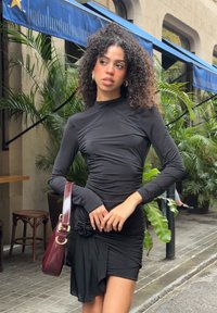 Fekete hosszú ujjú ruha ráncolt textúrával, virágos d�íszítéssel a deréknál. Bordó kézitáskával párosítva. Zöld növények a háttérben.