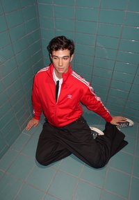 Rode Adidas-jas met ritssluiting en witte strepen, zwarte broek en marineblauwe sneakers. Zittend in een betegelde hoek met turquoise tegels.