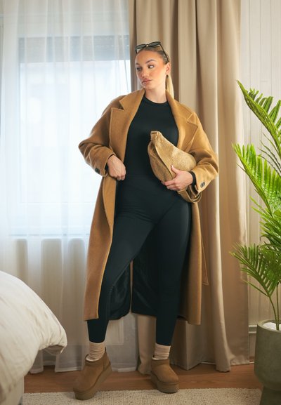 Abrigo de lana marrón sobre un bodysuit negro, leggings negros y botas plataforma color beige. Sosteniendo un bolso texturizado beige con forma curvada.