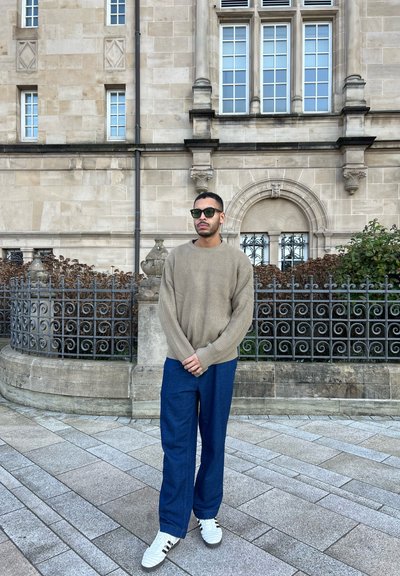 Suéter de canalé beige de gran tamaño, pantalones de pierna ancha azul oscuro y zapatillas blancas con acentos negros, todo ello frente a un edificio de piedra ornamentado.