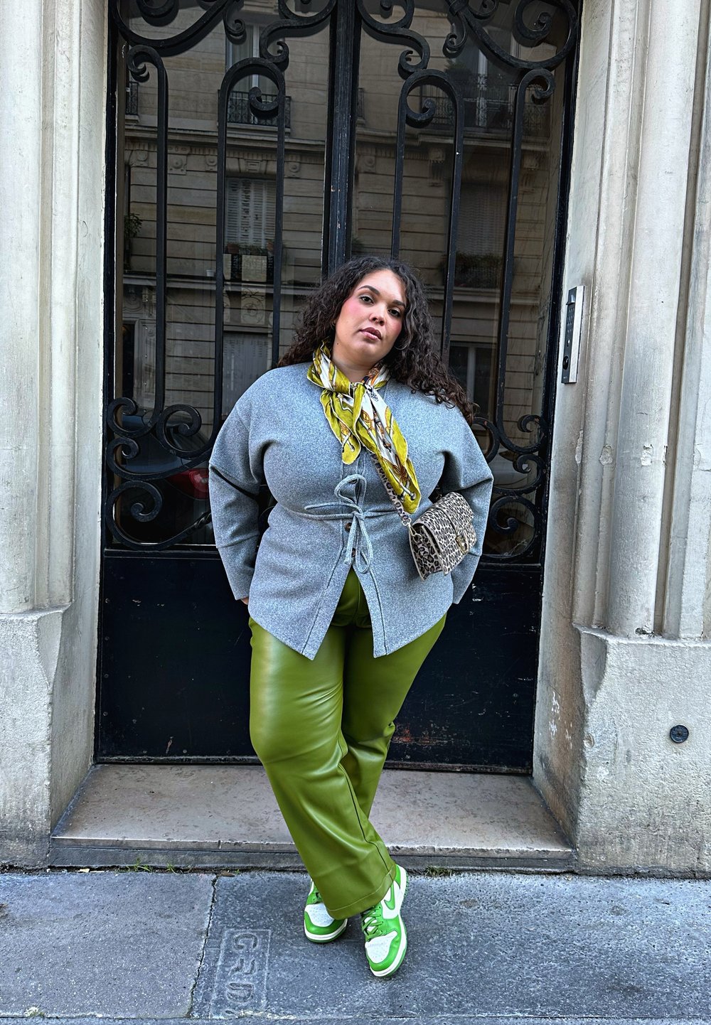 Grey knitted wrap cardigan with a tie waist, styled with green faux leather trousers and white trainers featuring green accents. Complete with a patterned scarf and handbag.