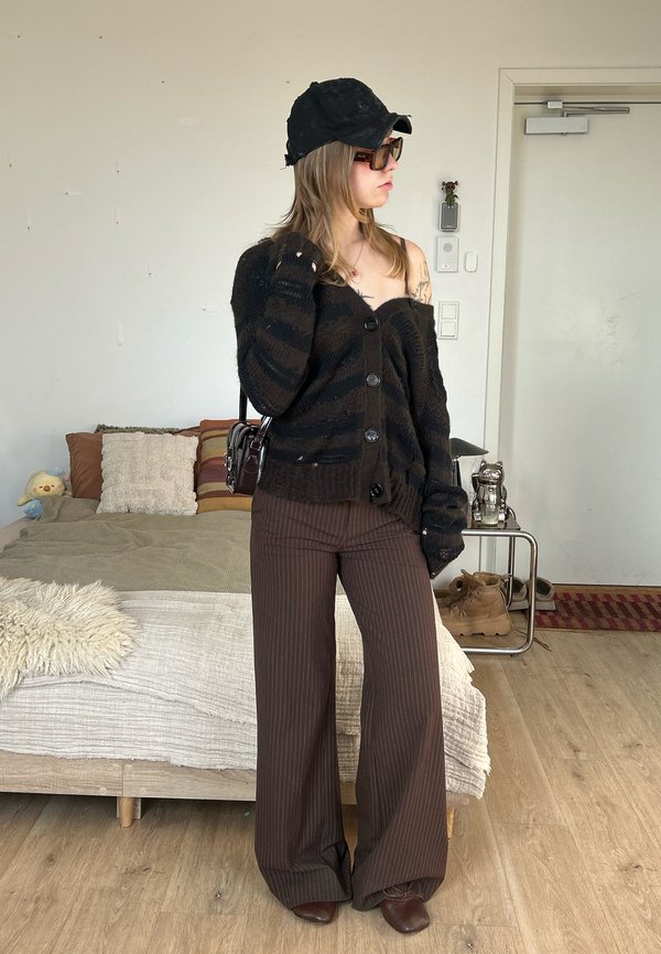 Ung kvinne iført svart caps, solbriller, oversized svart genser og brune vidbukser med pinstriper, stående i et koselig soverom.