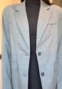 Blazer xadrez cinza com um corte ajustado, fecho de botão único, dois bolsos frontais e lapelas recortadas. Usado sobre um top preto.