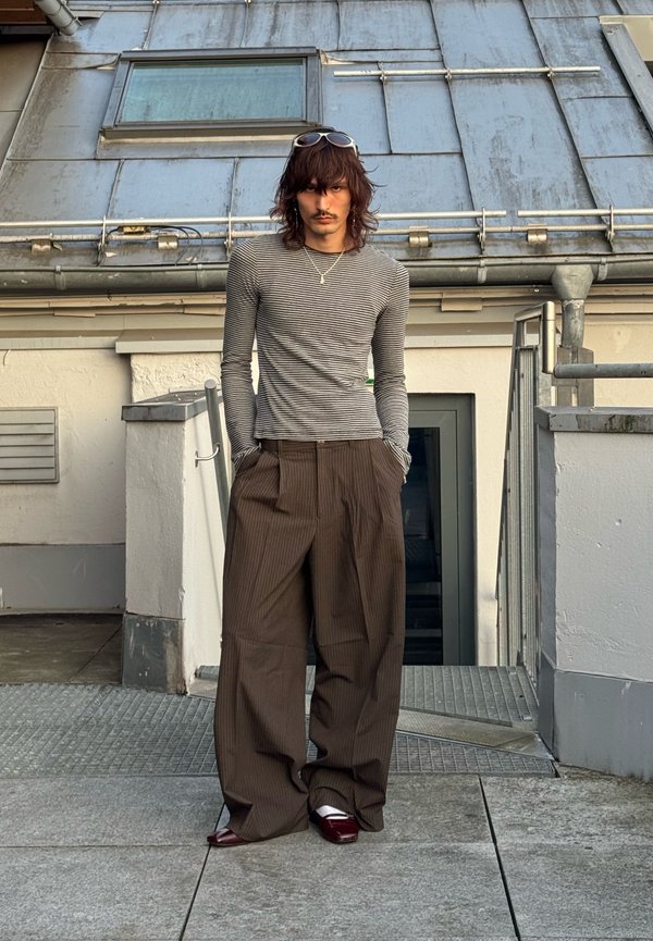 Homme aux cheveux mi-longs portant une chemise rayée à manches longues, un pantalon large marron et des chaussures bordeaux, debout sur les escaliers d'un toit urbain.