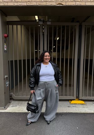 Mujer sonriendo, vestida con chaqueta de cuero negra, camiseta blanca, pantalones anchos grises, sosteniendo un bolso negro, de pie frente a una puerta metálica cerrada.