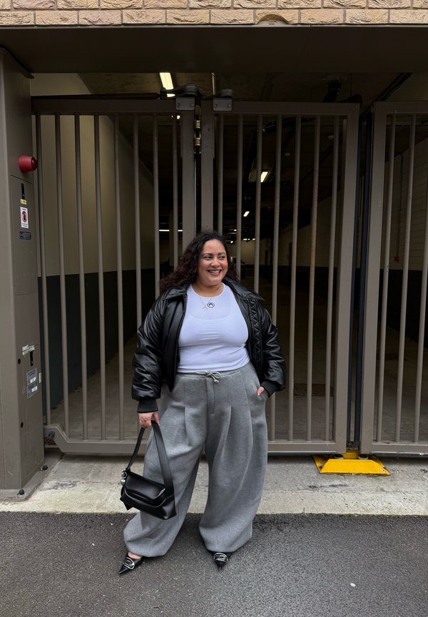 Mujer sonriendo, vestida con chaqueta de cuero negra, camiseta blanca, pantalones anchos grises, sosteniendo un bolso negro, de pie frente a una puerta metálica cerrada.