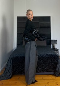 Jeune femme en haut noir et pantalon large gris tenant une grande pochette noire, debout à côté d'un lit avec une literie et une tête de lit sombres.