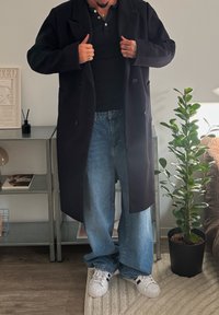 Personne ajustant un manteau noir, portant une chemise noire, un jean large bleu et des baskets blanches, debout près d'une étagère en métal et d'une plante verte en pot dans une pièce.