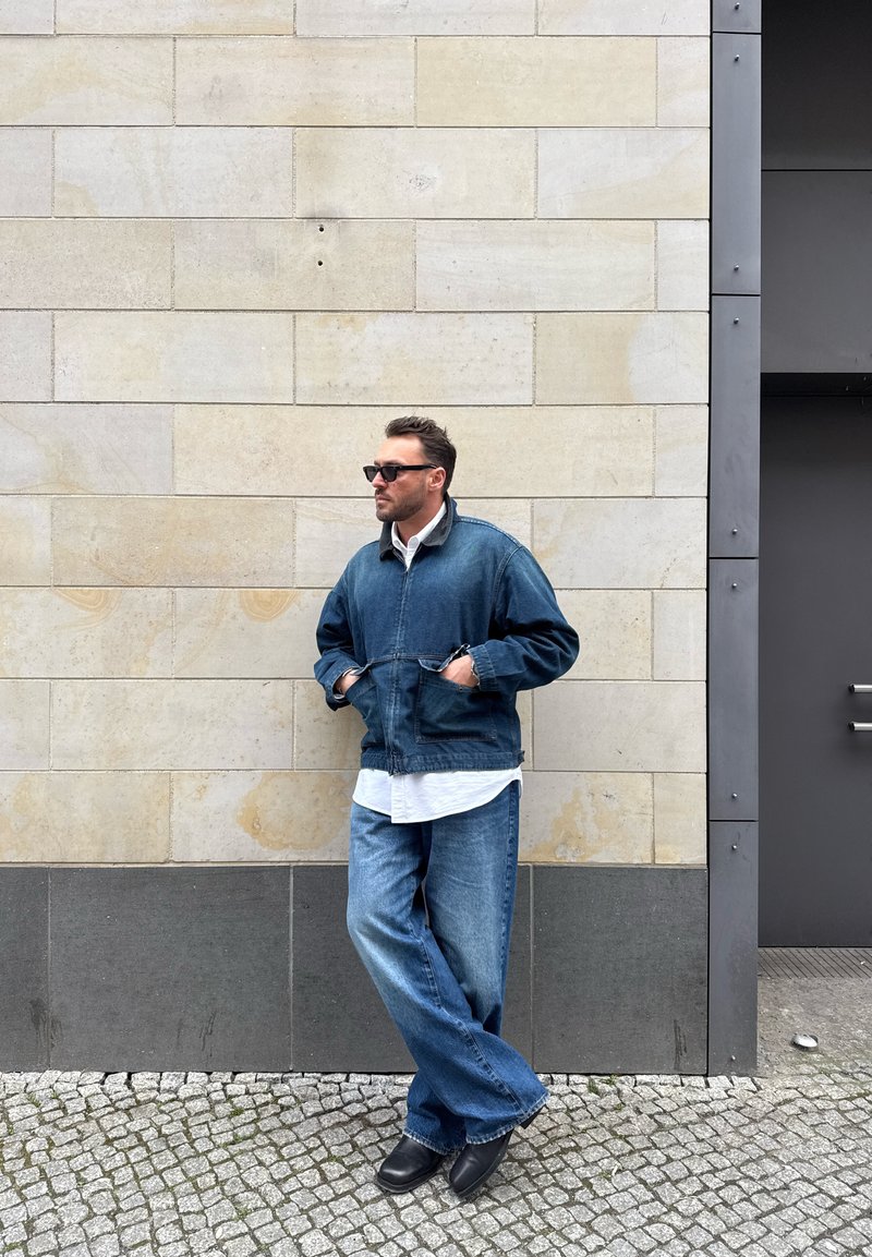 Homme portant des lunettes de soleil, une veste en jean, un jean ample et des chaussures noires, appuyé contre un mur de pierre claire sur un pavé en pierres.