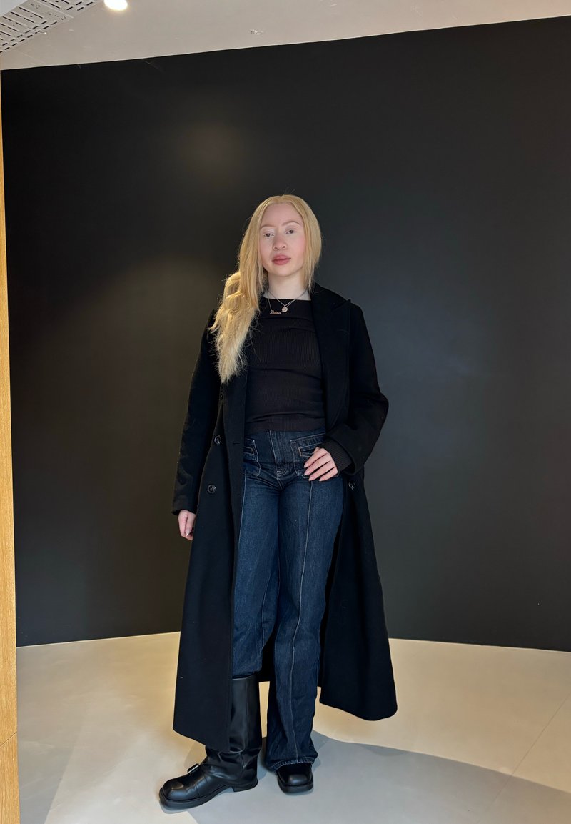 A black long coat worn over a black top, complemented by dark blue high-waisted jeans and black knee-high boots. Finished off with a simple silver necklace.