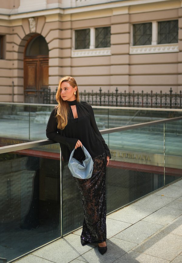Femme en blouse noire et jupe à sequins tenant un sac à main bleu clair, appuyée sur une rambarde en verre devant un bâtiment historique.