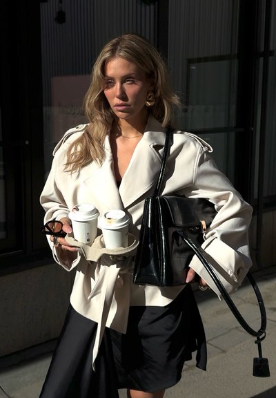 Mujer con abrigo tipo trench color crema y falda negra sosteniendo dos vasos de café para llevar y un bolso grande negro mientras está de pie al aire libre.