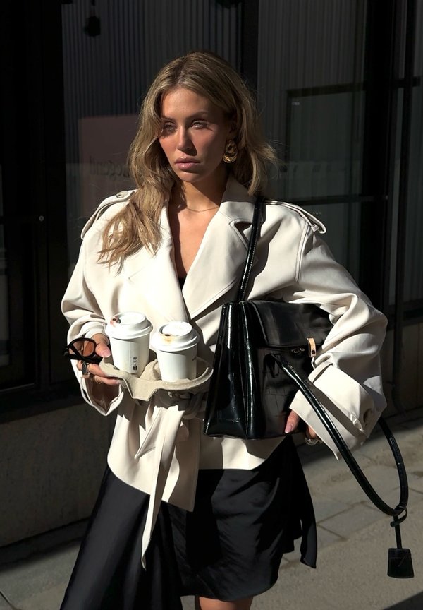 Vrouw in een crèmekleurige trenchcoat en een zwarte rok die twee koffie-to-go bekers en een grote zwarte handtas vasthoudt terwijl ze buiten staat.