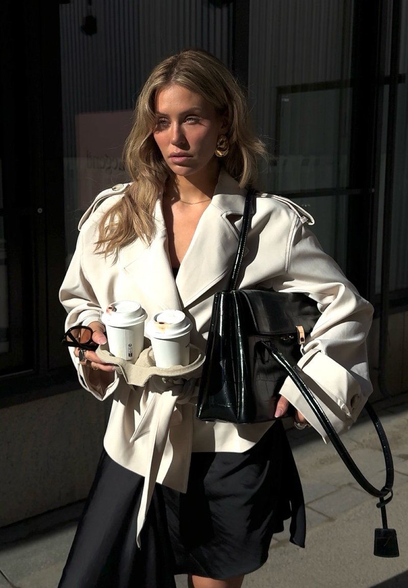 Kvinde iført cremefarvet trenchcoat og sort nederdel, der holder to takeawaykaffekopper og en stor sort håndtaske, mens hun står udendørs.