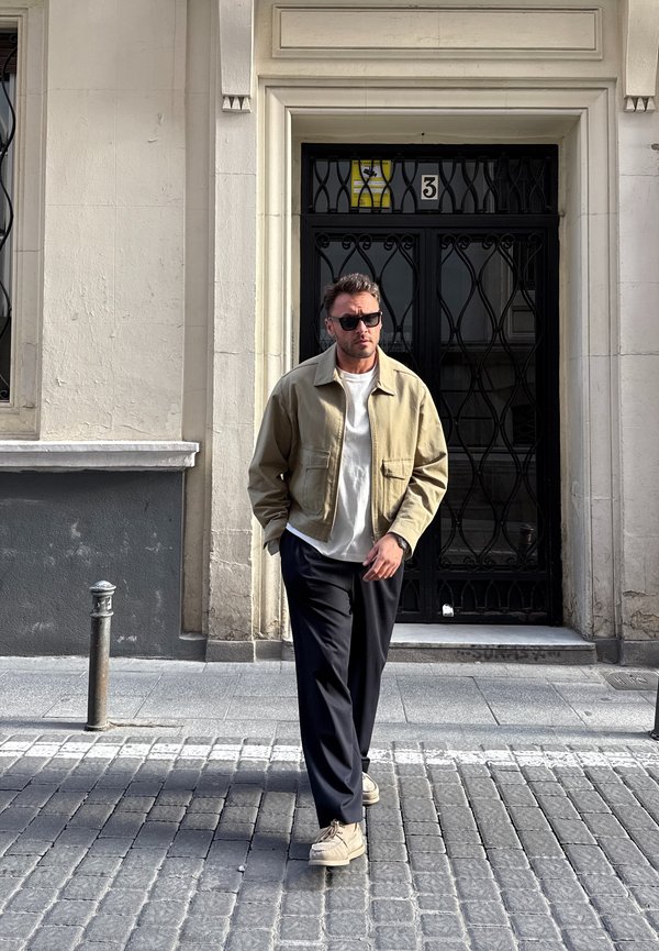 Άνδρας με γυαλιά ηλίου, μπεζ σακάκι, λευκό πουκάμισο και μαύρο παντελόνι περπατάει σε λιθόστρωτο δρόμο μπροστά από μαύρη μεταλλική πόρτα με αριθμό 3.