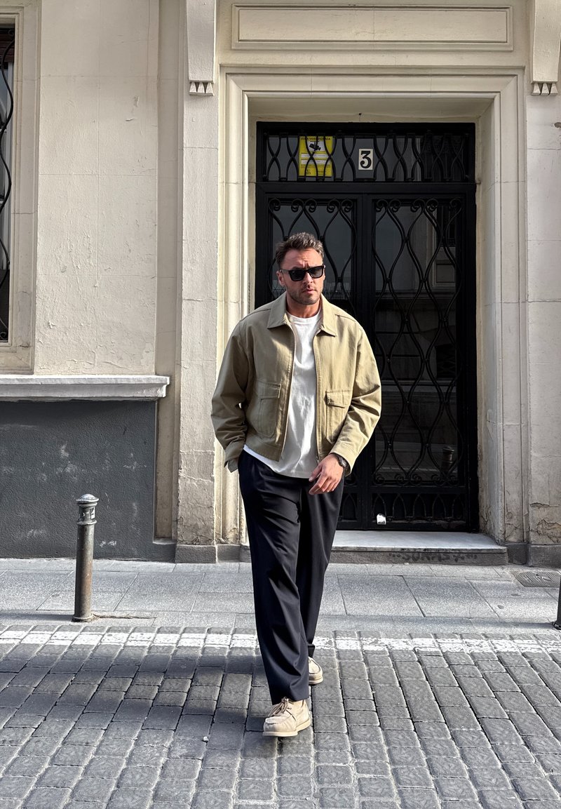 Homme portant des lunettes de soleil, une veste beige, une chemise blanche et un pantalon noir marchant sur une rue pavée devant une porte métallique noire avec le numéro 3.