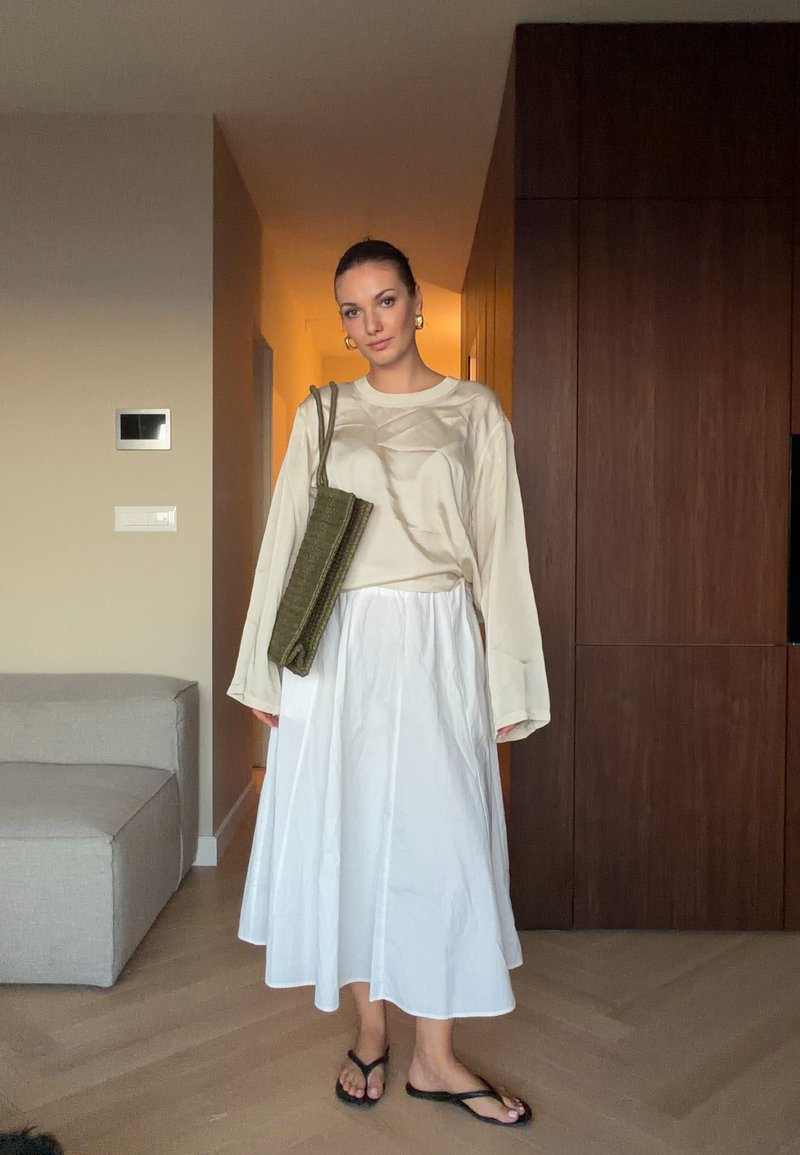 Femme portant un haut beige à manches longues, une jupe midi blanche, des sandales noires et portant un sac à épaule vert olive dans un salon moderne.