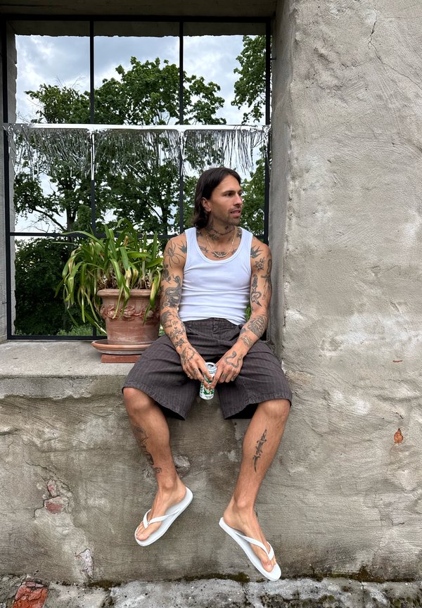 Uomo con tatuaggi che indossa canotta bianca, pantaloncini neri e infradito bianchi seduto su un bordo di cemento tenendo una lattina, con pianta in vaso e alberi dietro.