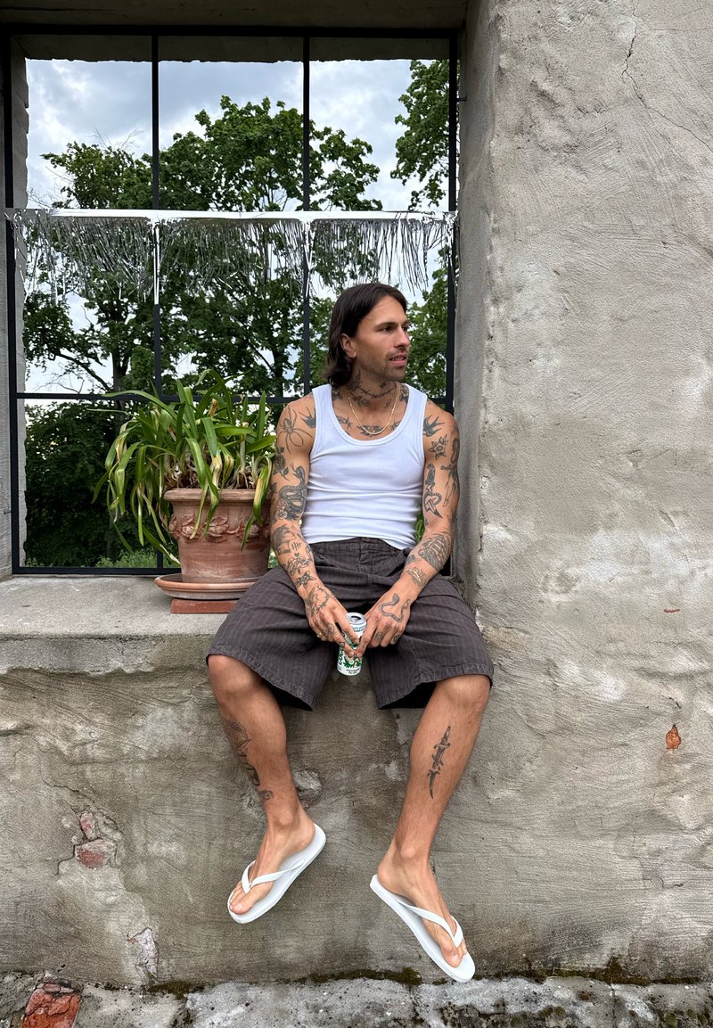 Homme tatoué portant un débardeur blanc, un short noir et des tongs blanches, assis sur un rebord en béton tenant une canette, avec une plante en pot et des arbres derrière.