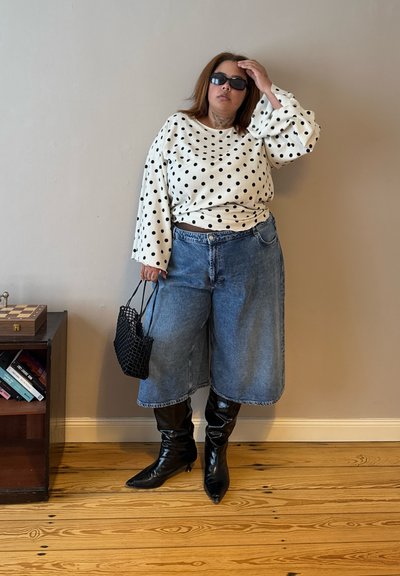 Mujer con gafas de sol negras, blusa blanca de lunares, pantalones cortos anchos de mezclilla, botas negras, sosteniendo una bolsa negra de red, de pie en el interior junto a una estantería.