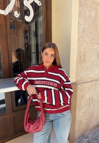 Rood en wit gestreepte gebreide trui met een kraag, voorzien van de tekst "EMESTUDIOS", gecombineerd met een bordeauxrode geweven handtas en lichtblauwe jeans.